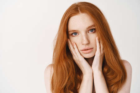 Beauty and skincare. Close-up of redhead pale woman touching face, looking at camera sensual, showing perfect no makeup skin, standing over white backgroundの写真素材