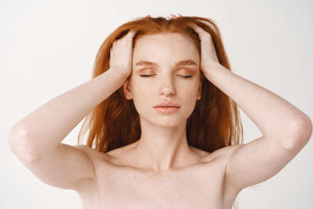 Close-up of relaxed young redhead woman with pale skin and freckles, massaging natural red hair with...