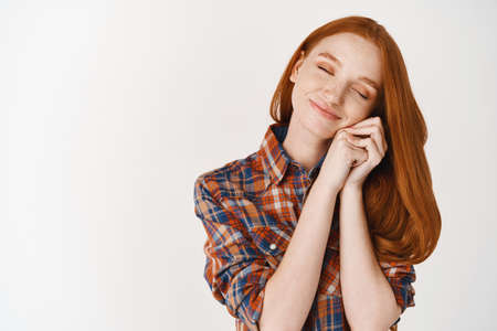 Lovely and romantic redhead girl dreaming, close eyes and leaning on hands tenderly, standing over white background and smilingの写真素材