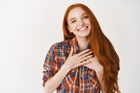 Cute redhead woman with pale skin and blue eyes, holding hands on heart and smiling thankful, express gratitude and happiness, standing over white backgroundの写真素材