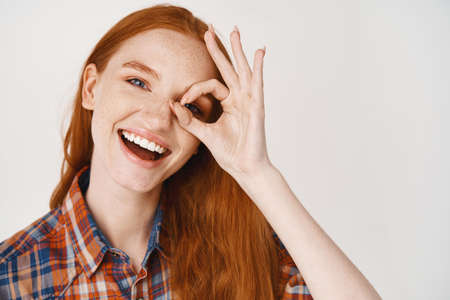 Close-up of happy redhead woman with pale skin and white teeth, showing okay sign on eye and...