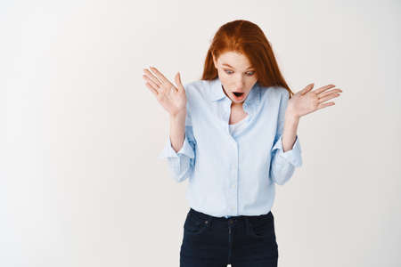 Surprised redhead girl staring down at promo special deal, gasping and spread hands sideways amazed,...