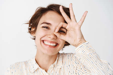 Close up of natural young woman showing okay sign on eye and smiling, approve something,...