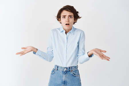 Confused and anxious young woman shrugging shoulders, spread hands sideways and looking puzzled at camera, know nothing, standing on white backgroundの写真素材