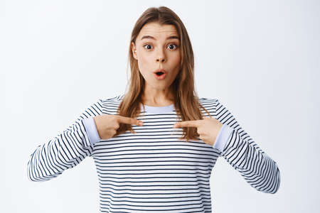 Surprised and excited blond woman pointing at herself and look with disbelief at camera, being chosen or picked as winner, standing in casual outfit over white backgroundの写真素材