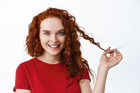 Portrait of confident beautiful ginger girl with curly hairstyle, playing with curl strand and smiling sassy at camera, staring intrigued, standing in t-shirt against white backgroundの写真素材