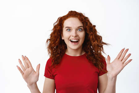 Portrait of surprised ginger girl with curly hair, raise hands and gasping amazed, checking out awesome advertisement. Redhead woman screaming excited about surpriseの写真素材