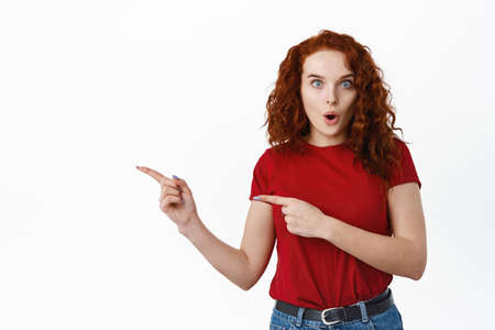 Impressed redhead girl gasping, saying wow as staring at camera amazed and pointing fingers left at exciting promo deal, showing advertisement, white backgroundの写真素材