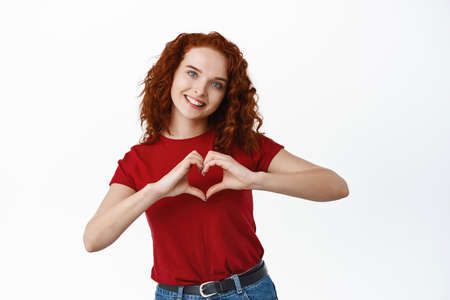 Portrait of beautiful ginger girl with curly hair and pale skin, showing heart I love you gesture...