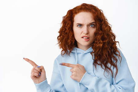 Portrait of confused woman with red curly hair staring awkward and puzzled at camera, biting lip and frowning, pointing fingers left at copy space logo, white backgroundの写真素材