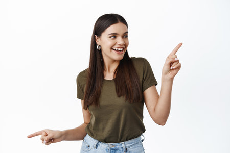 Young millennial girl with joyful smile going on shopping, facing choice, pointing sideways and picking product in store, having variants, white backgroundの写真素材