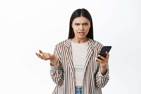 Portrait of confused and annoyed girl frowning and grimacing after seeing something strange on phone, holding smartphone and shrugging, white backgroundの写真素材