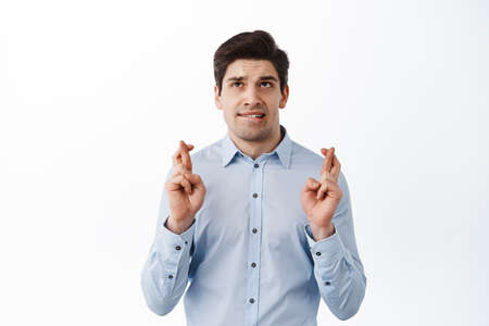Nervous office worker praying, cross fingers for good luck and looking up hopeful, begging, anticipating good news with worried face, white backgroundの写真素材