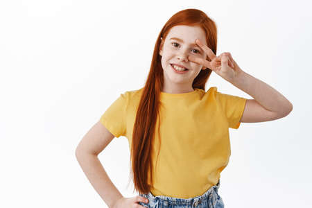 Positive little girl with long ginger hair and freckles smiling happy, showing peace kawaii sign on eye, posing in yellow t-shirt against white backgroundの写真素材
