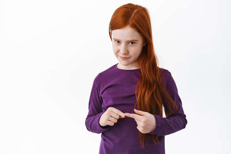 Shy little ginger girl, kid with red hair looking modest touching two fingers and looking from under...