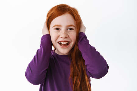 Cute little kid with red hair shut her ears and smiling, acting childish, unwilling to listen, misbehave, laughing and having fun, standing over white backgroundの写真素材