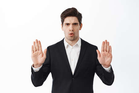 No, stop this. Serious corporate man, businessman in suit frowning, blocking offer, rejecting, making decline refuse gesture, standing disappointed against white backgroundの写真素材