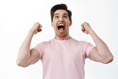 Success and celebration. Excited man looking up at screen, winning bet, shaking hands and scream yes, celebrating victory, win and triumphing, standing over white backgroundの写真素材
