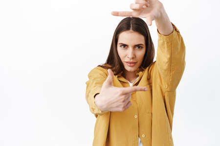 Creative and passionate woman searching perfect angle, looking through hand frames camera gesture and determined pensive look, creating something, standing over white backgroundの写真素材