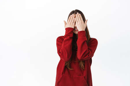 Cute caucasian woman in red hoodie covering her face and eyes with palms, smiling, waiting for...