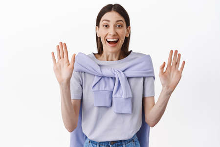 Happy pretty female model raising hands up, showing empty palms and looking excited at camera,...