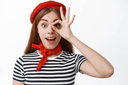 Cheerful young woman in french red beret and striped t-shirt, look through okay sign, make OK gesture over eye and gasping excited, white backgroundの写真素材