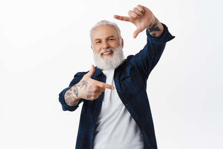 Smiling senior man makes hand frames camera gesture, taking picture and smiling, making snap while...