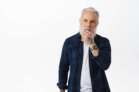 Thoughtful hipster old man touch his beard and looking at upper left corner, pondering choice,...