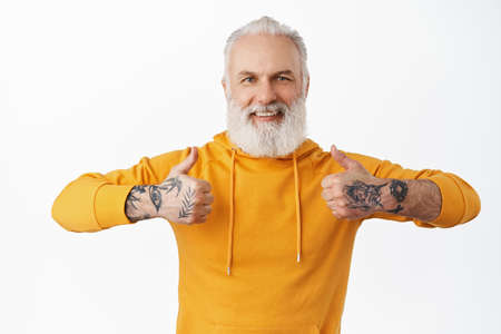 Supportive handsome senior man with tattoos, grandfather showing thumbs up in approval, agree and like something good, praise nice choice, agree and say yes, white backgroundの写真素材