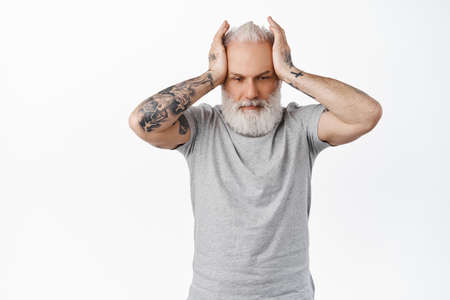 Annoyed and distressed old man with tattoos, holding hands on head with troubled upset face, looking...