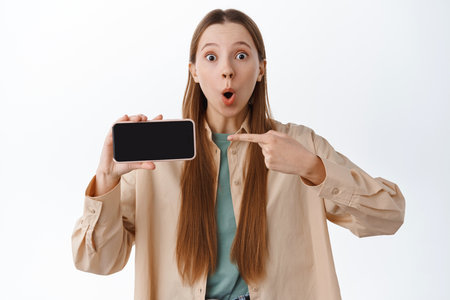 Surprised millennial girl say wow, pointing at horizontal smartphone screen, showing mobile display with amazed face, standing over white backgroundの写真素材