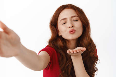 Romantic redhead woman sending air kiss with cute flirty face, taking selfie or video calling lover,...