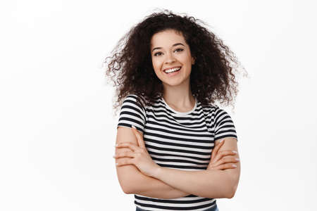 Image of brunette curly girl smiling confident, cross arms on chest, looking pleased at camera,...