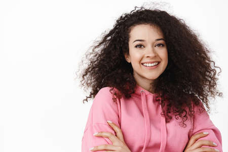Close up portrait of modern brunette girl with natural curly hair, cross arms on chest, smiling white teeth, looking happy and relaxed at camera, standing against white backgroundの写真素材