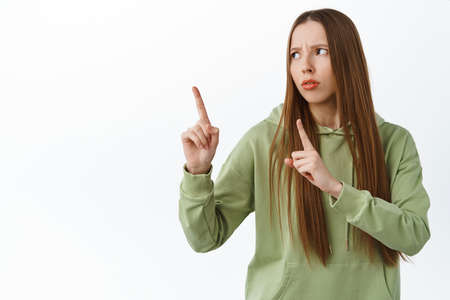 Confused teenage girl with long hair, pointing and looking aside at left copyspace, frowning...