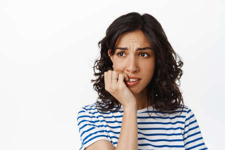 Close up of anxious brunette girl, biting finger nails and looking away with alarmed, nervous face,...