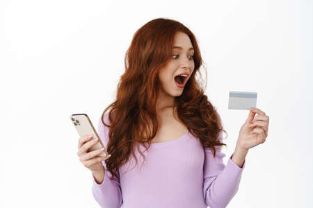 Online shopping. Redhead girl looks amazed at credit card, order something in internet, holding...