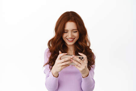 Smiling redhead woman watching video on smartphone screen, looking at mobile phone happy, playing...