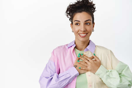 Young lesbian woman holding hand on heart, smiling happy and looking away at logo, being uplifted,...