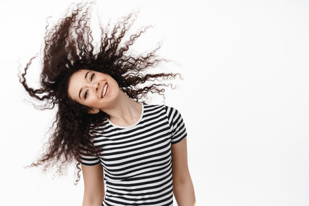 Portrait of happy natural brunette woman flip curly hair and smiling carefree, looking cheerful, standing against white background and having funの写真素材