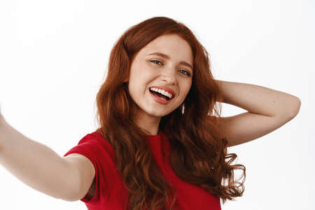 Close up portrait of ginger girl taking selfie on smartphone, posing at camera, laughing and smiling happy, standing in t-shirt against white backgroundの写真素材