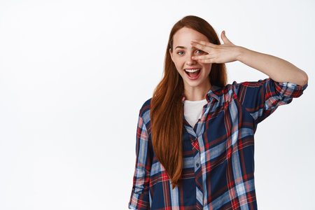 Happy positive teen girl with red hair, laughing and smiling, open eyes, peeking through fingers and laughing natural, standing against white backgroundの写真素材