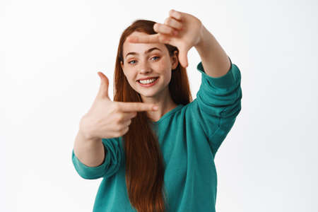 People and lifestyle. Happy redhead woman look through hand frames and smiling, imaging and getting creative, standing over white backgroundの写真素材