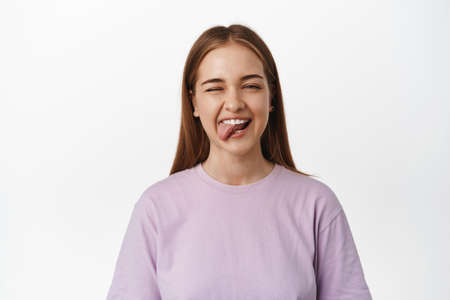 Close up of positive and carefree girl shows tongue and winks, feel happy and upbeat on summertime holidays, enjoying event, standing against white backgroundの写真素材