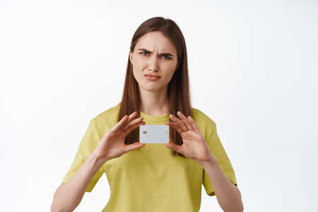 Angry and displeased girl shows credit card, frowns and grimace upset, complain about bank or lack...