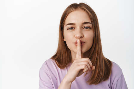 Hush. Close up portrait of blond girl shushing, smiling and asking to keep quiet, has secret, shhh...