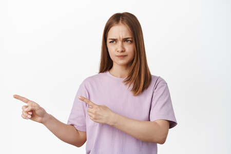 Displeased gloomy girl furrow eyebrows, look and pointing left at bad, unfair or disappointing,...