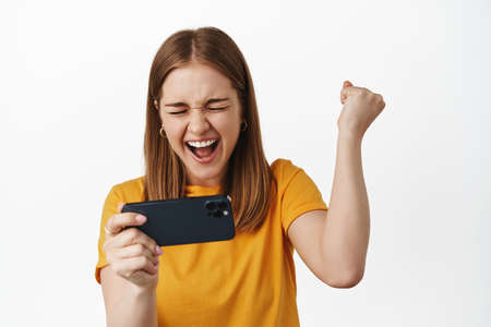 Blond woman winning money on phone. Girl rejoice, holding smartphone horizontal, playing mobile...