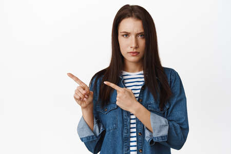 Disappointed frowning woman pointing left, looking serious and reluctant, dislike promotion, so low discount, waiting for explanation, standing upset against white backgroundの写真素材