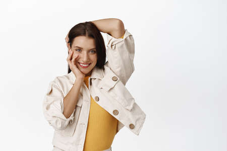 Portrait of attractive sassy woman, coquettish daring smile, touching her head, hair and face, posing flirty in stylish outfit, standing against white backgroundの写真素材
